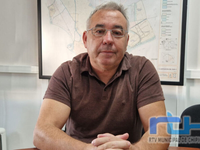 Un hombre con cabello gris y gafas de sol, vestido con una camiseta marrón, sentado en un escritorio frente a un mapa de la ciudad. En el fondo se puede ver una pared con paneles de información y una señalética de la RTV Municipade Chipiona.