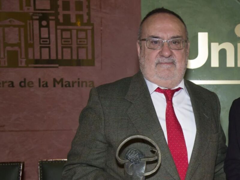 Un hombre en traje y corbata roja, posiblemente un político o funcionario público, frente a una bandera de la Unión Europea y el Ministerio de Defensa.