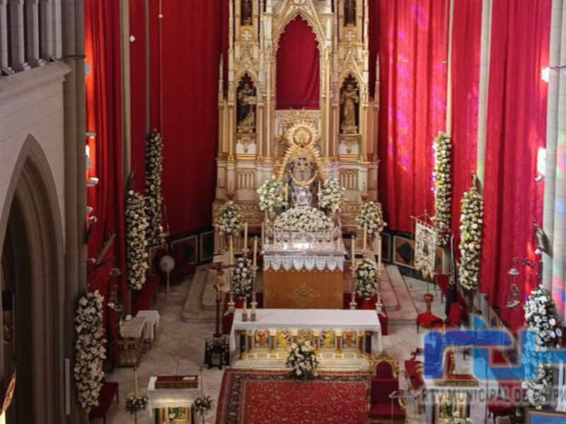Interior de una iglesia con altares dorados y flores blancas, rodeado por cortinas rojas.
