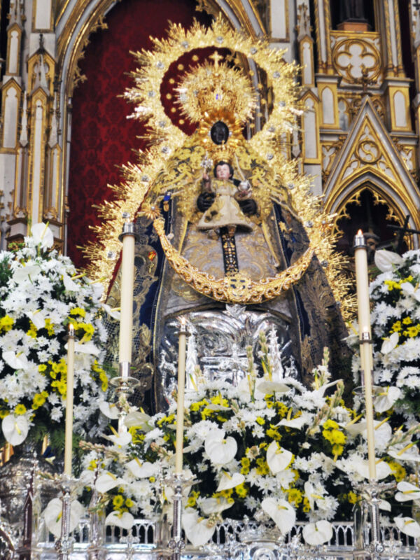 Imagen de una imagen religiosa con flores blancas y amarillas en un altar, destacando la figura central de una Virgen con un niño.