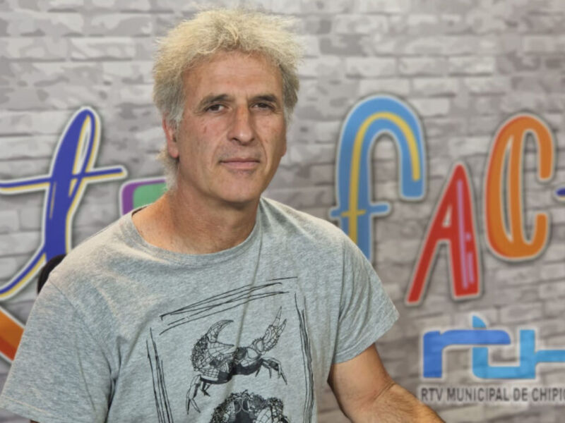 Un hombre con cabello canoso y una camiseta gris posa frente a un fondo con letras coloridas.