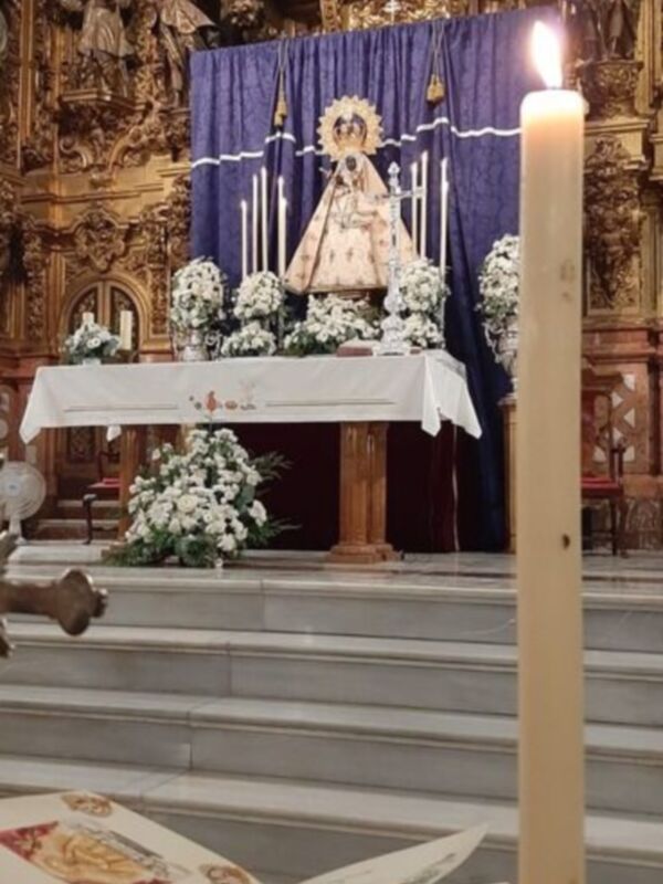 Una imagen de una iglesia con un altar adornado con flores y velas encendidas. En el centro, una estatua de la Virgen María rodeada por flores y velas. En el fondo, detalles arquitectónicos elaborados.