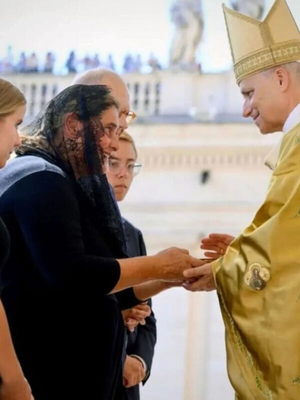 Una persona en traje negro y tocado dorado estrecha la mano de otra persona con un atuendo formal. Fondo blanco con detalles blancos y dorados.