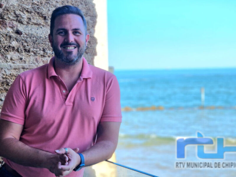 Un hombre con una camiseta rosa posa frente a un edificio de piedra, con el mar y el cielo azul en el fondo. La imagen está firmada por RTV Municipal de Chipiona.