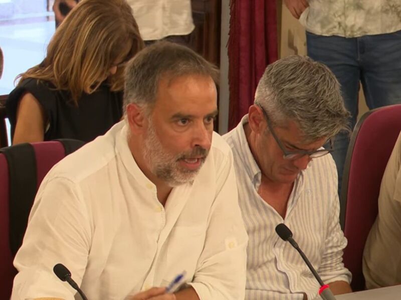 Un grupo de personas se reúne en una sala de conferencias. La imagen muestra a varios individuos sentados, uno de ellos con un micrófono en frente. La sala tiene sillas rojas y una pared con cortinas de color rojo. Algunos detalles como la ropa casual, el micrófono y los asistentes sugieren una reunión formal o un evento de negocios.