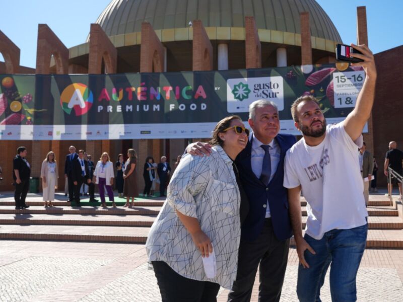 Fotografía de una familia tomando un selfie frente a un edificio con un cartel que dice "Auténtica Premium Food" y una fecha de 15-16. El ambiente es soleado, con personas vestidas formalmente y otros en traje casual.