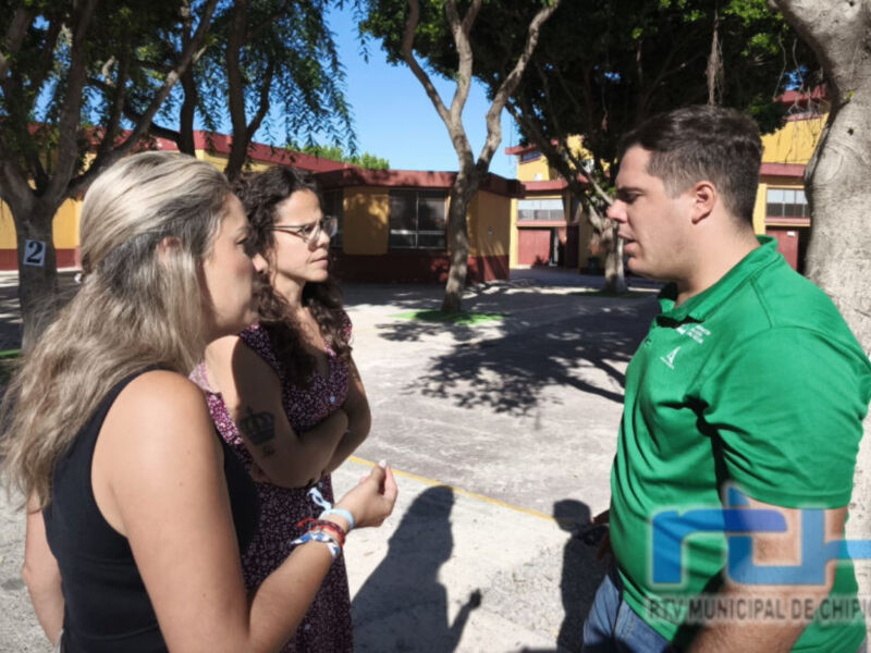 Tres personas conversan en una calle con árboles y edificios al fondo. La imagen muestra un ambiente urbano tranquilo con elementos de la ciudad en el horizonte.