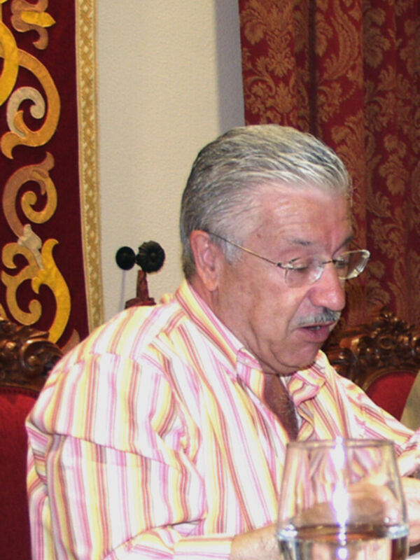 Un hombre con cabello gris y barba, vistiendo una camisa de rayas, se sienta en un ambiente formal con cortinas rojas y doradas. En la mesa frente a él hay una copa de vino.