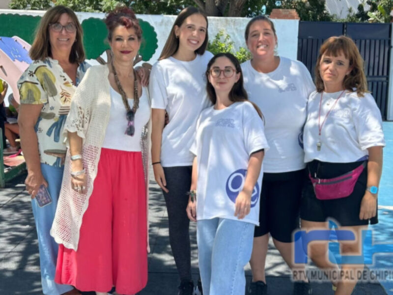 Una foto de cinco personas posando en una calle, con un fondo de pared blanca y cielo azul. La imagen parece ser parte de una celebración o evento público, con algunos asistentes en el fondo. La foto es cortada y no muestra la totalidad de los individuos o el entorno completo.