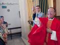 Un sacerdote en traje rojo y corbata celebra con un gesto de la mano, mientras una persona en el fondo toma una foto. La imagen se sitúa en un interior con puertas doradas y vidrieras, y una placa de salida visible.