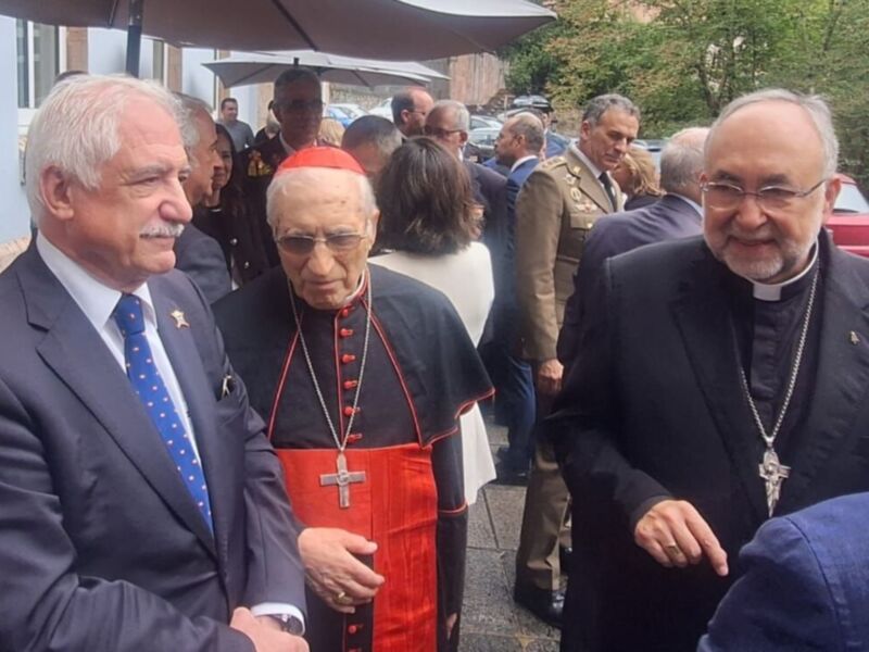 Se observa a varios hombres, algunos de ellos con trajes y otros con vestimentas religiosas. La escena parece ser una reunión o evento formal, posiblemente en un entorno público.