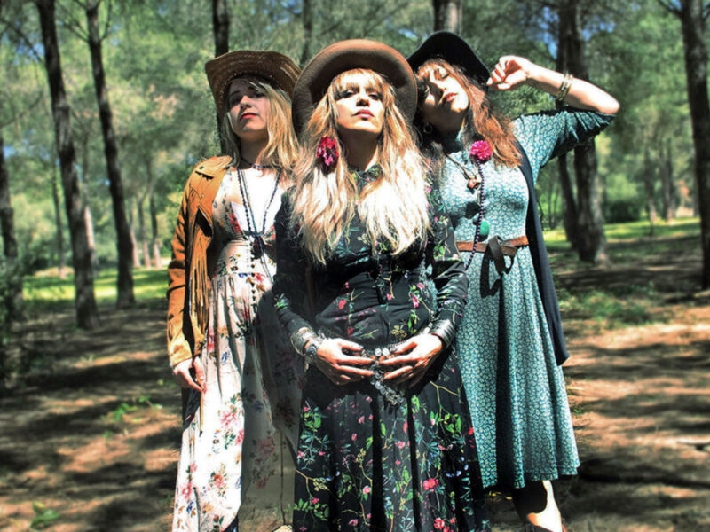 Tres jóvenes en un bosque, vestidas con sombreros y vestidos de colores vibrantes. La escena es soleada, con árboles y vegetación alrededor.