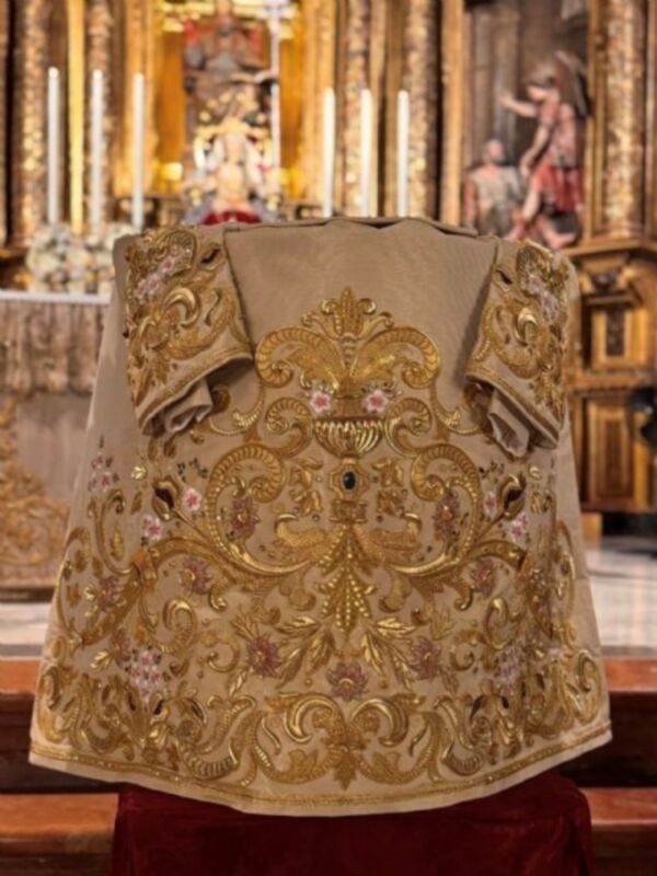 Elegante chasuble bordado en dorado y oro con detalles florales, frente a un altar de madera tallada.