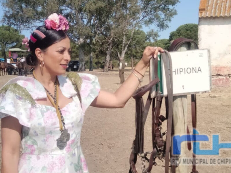 Chica con vestido floral y tocado de flores, sosteniendo un signo que dice "Chipiona". Fondo con árboles y estructuras de tierra.