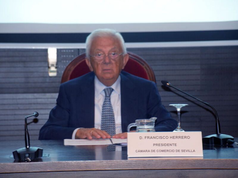 D. Francisco Herrero, Presidente de la Camara de Comercio de Sevilla, sentado en una mesa con un nombreplatea y dos micrófonos.