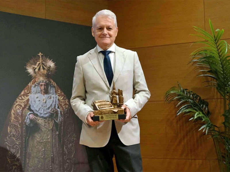 Un hombre en traje blanco sostiene un trofeo dorado con una estatua de la Virgen María en el fondo. La imagen está ambientada en un entorno formal con una pared de madera y una planta verde a la derecha.