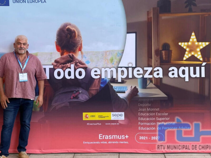 Un hombre con una camiseta rosa y un lanyard está frente a un póster de la Unión Europea que muestra una persona sentada en un escritorio con un portátil y un mapa. El póster tiene texto que dice "Todo empieza aquí" y menciona Erasmus+ 2021-2022.