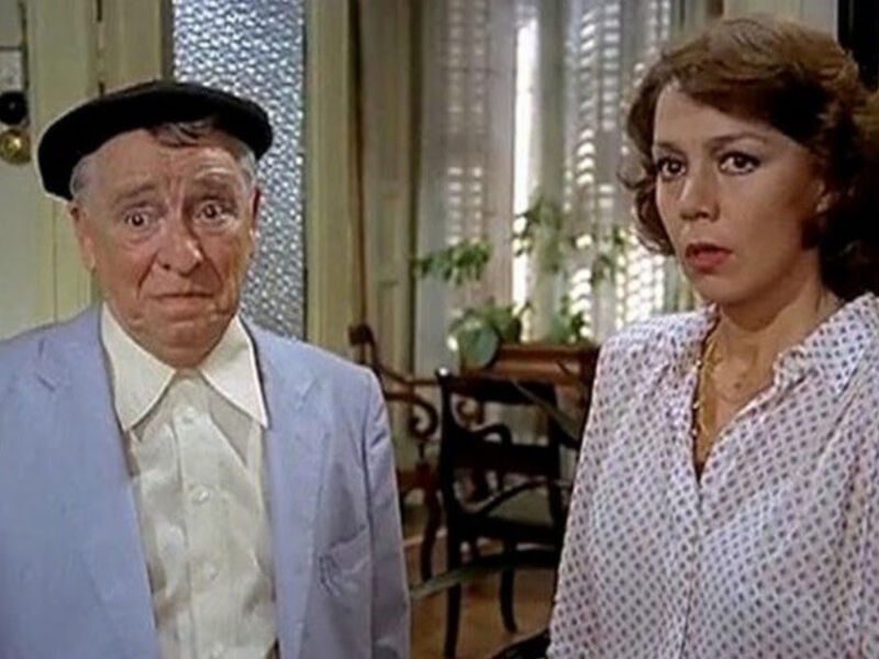 Dos personajes en una escena de película, uno con un traje azul y el otro con una camisa blanca a cuadros. Ambos están en un ambiente interior, posiblemente una sala de estar o comedor, con decoración clásica y un piano visible en el fondo.