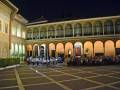 Un patio de noche con una arquitectura histórica, gente sentada en el suelo y un grupo de músicos tocando al fondo.
