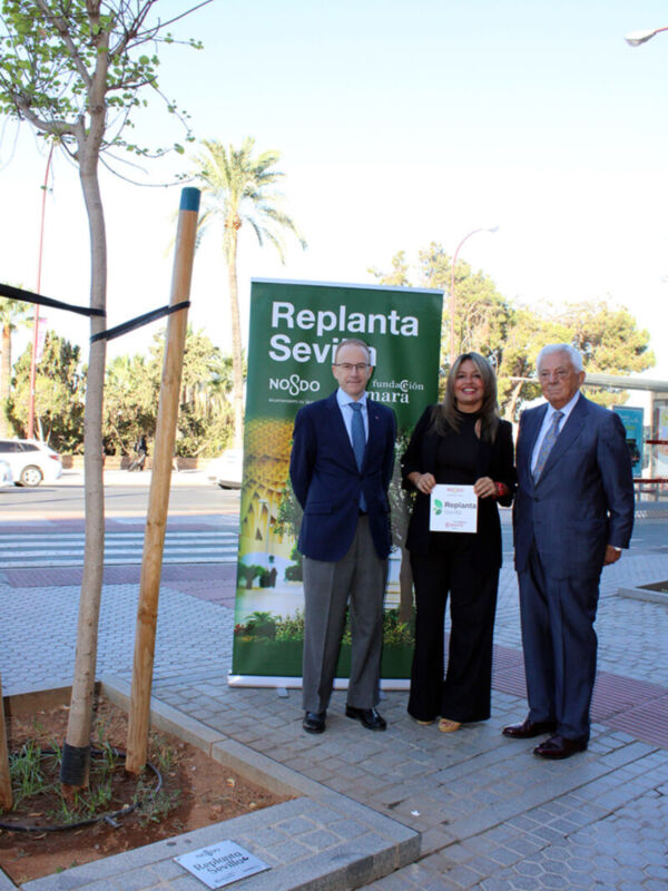 A tres personas posando frente a un cartel que anuncia "Replanta Sevilla" y "Noox". El cartel está ubicado en una calle con árboles y edificios al fondo. La escena parece ser parte de una iniciativa o evento relacionado con la reforestación en Sevilla.