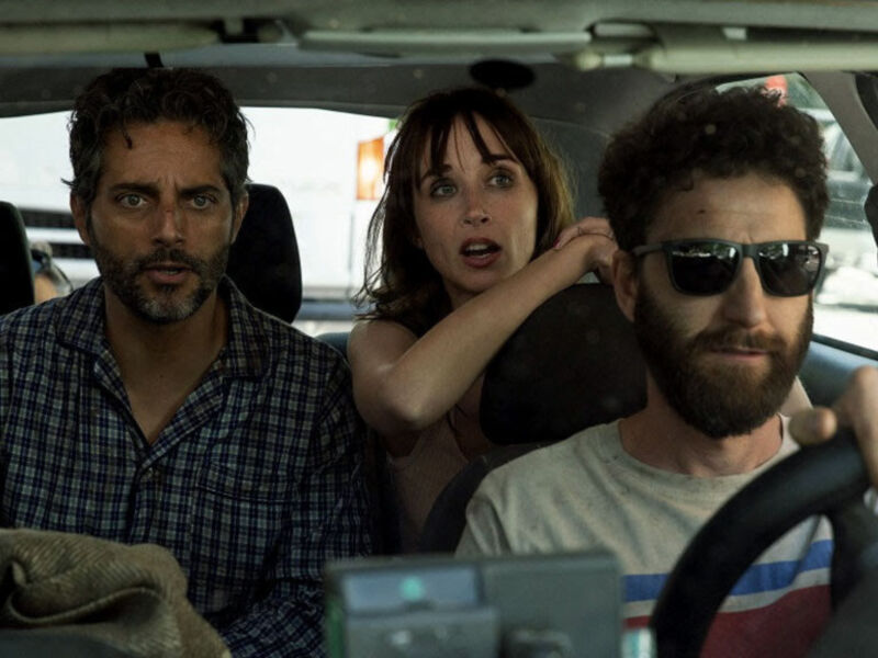 Un hombre con barba y camisa a cuadros está conduciendo un coche, mientras una mujer con cabello largo y una persona más joven con gafas de sol se sientan en el asiento del pasajero. La imagen parece ser una escena de un filme o cortometraje.