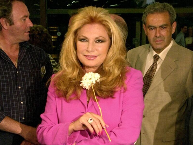 A una mujer con cabello largo y rubio, vestida de rosa, sosteniendo un ramo de flores blancas. A su lado están dos hombres, uno con una camisa a cuadros y el otro con un traje beige. La escena parece ser en una sala de conferencias o evento formal.