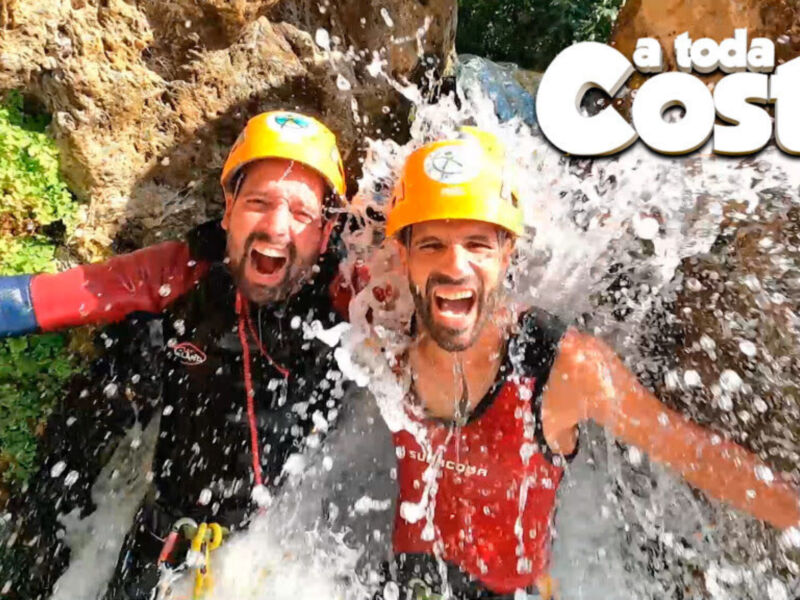 Dos personas en cascos de seguridad disfrutan de un descenso en cascadas, con agua salpicando y rocas verdes alrededor. Texto: "a toda Costa" en la parte superior derecha.