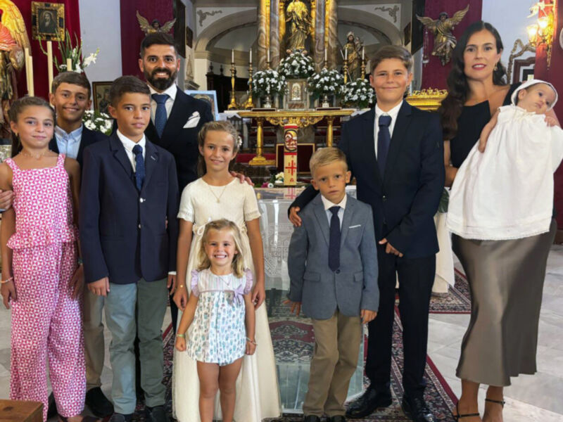 Fotografía de una familia numerosa en un ambiente religioso, con niños vestidos formalmente y adultos sonrientes. La imagen se toma en un entorno decorado con elementos religiosos, como estatuas y velas.