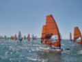 Una imagen de vela windsurf en un día soleado con el mar azul y la brisa soplando. En primer plano, una embarcación con un velo naranja y la bandera española. En segundo plano, varios otros windsurfers con velas de colores diferentes. El cielo es claro y brillante, reflejando en el agua.