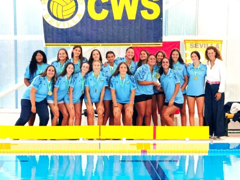 Equipo femenino de natación posando frente a una piscina con banderas del CWS y el logo de Sevilla.
