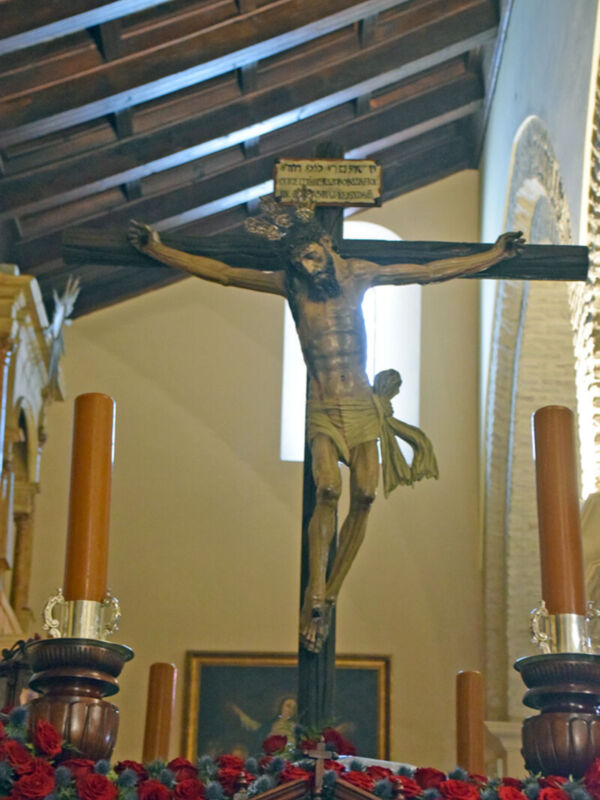 Crucifijo en una iglesia con detalles de madera y velas encendidas. Arriba, techo de vigas de madera. Abajo, flores rojas y un cuadro en el fondo.