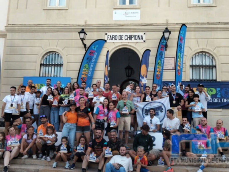 A una gran cantidad de personas posando juntas frente a un edificio con el nombre "TARO DE CHIPIONA" en la parte superior. La mayoría de las personas están sosteniendo carteles y sonríen para la foto. El ambiente parece ser festivo, posiblemente relacionado con un evento deportivo o social.