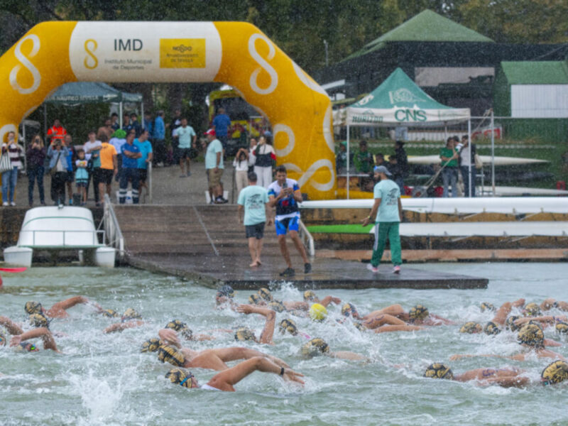 Competencia de natación en un estanque, con nadadores en acción y una gran bandera amarilla de la empresa IMD al fondo.