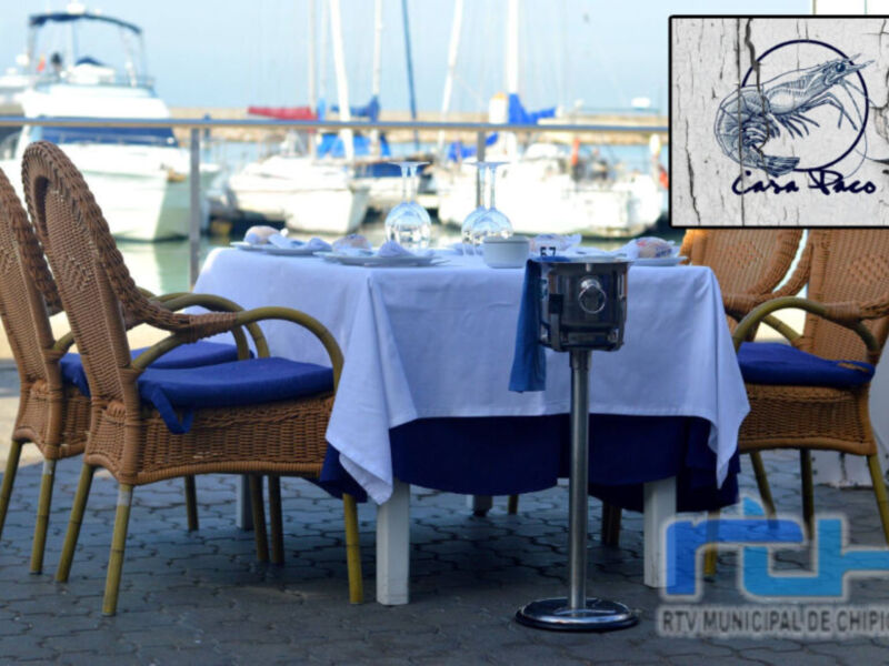 Silla de madera con cojín azul, mesa con mantel blanco y azul, fondo de embarcaciones en el puerto.