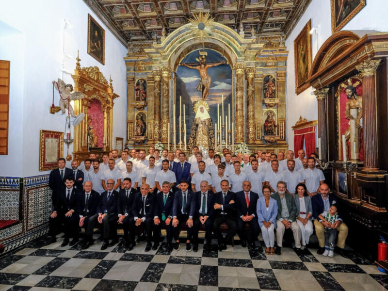 Grupo de personas posando en una iglesia con un altar dorado y una imagen del Cristo crucificado. La decoración es elaborada, con estatuas y paneles artísticos en el fondo. La imagen muestra una reunión formal o religiosa.