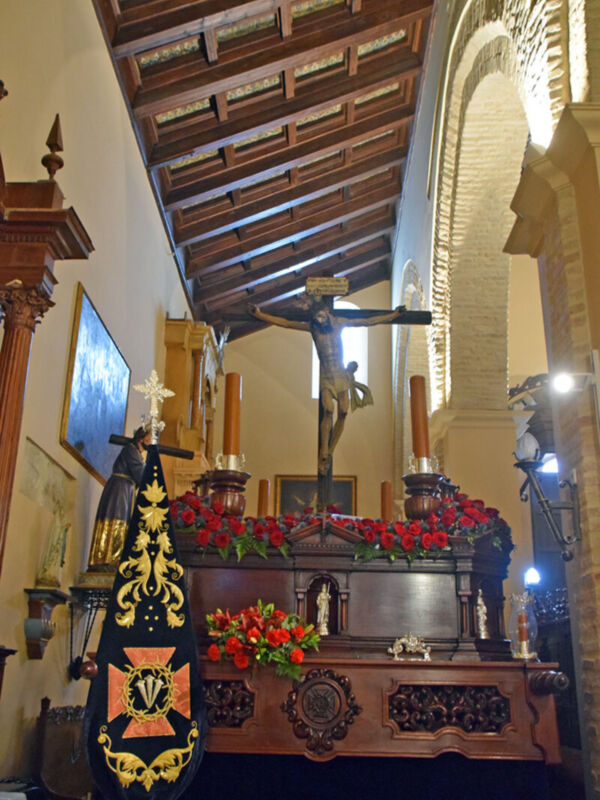 Interior de una iglesia con un altar principal, flores rojas en el centro y estatuas religiosas a ambos lados. Arriba, una escalera con detalles de madera y un techo de vigas.