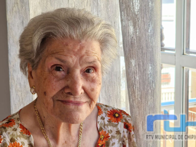 Una anciana con cabello gris y una camisa floral, posando frente a una puerta de madera.