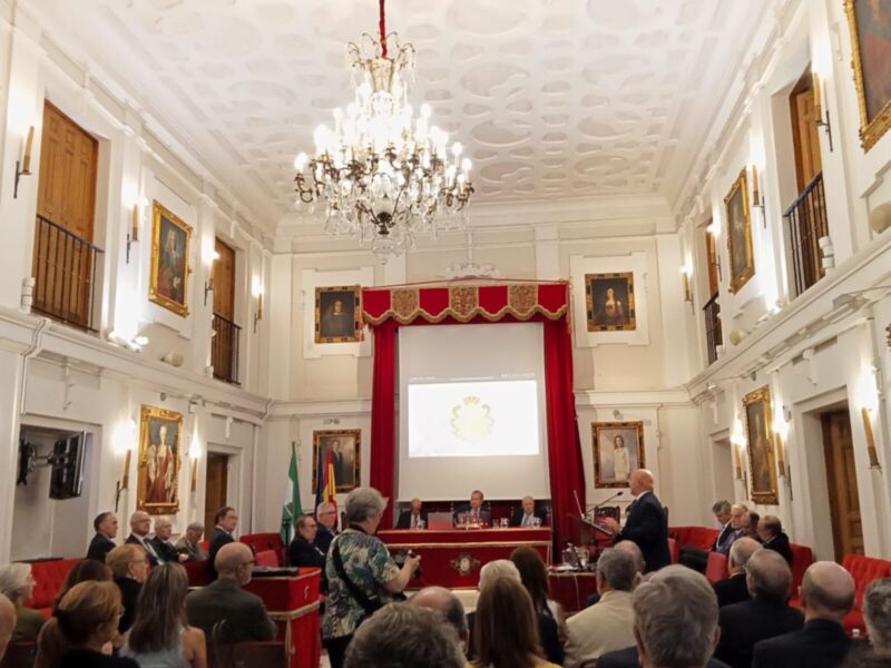 Una sala de conferencias con una iluminación elegante, un lustre en el techo y varias imágenes de retratos. La gente está sentada en sillas rojas, con un hombre hablando frente a una pantalla proyectora. La decoración es formal y sofisticada, con detalles dorados en el techo.