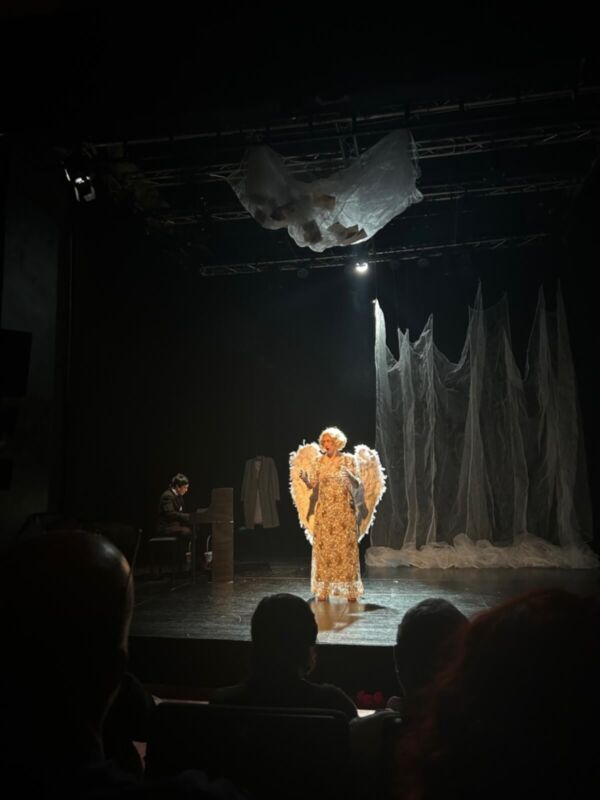Una figura vestida de dorado con alas gigantes y plumas en la escena teatral, frente a un público que observa atentamente. La iluminación enfoca la figura en el centro del escenario, rodeado por una atmósfera misteriosa con un fondo de tela negra y un objeto en forma de globo colgando del techo.