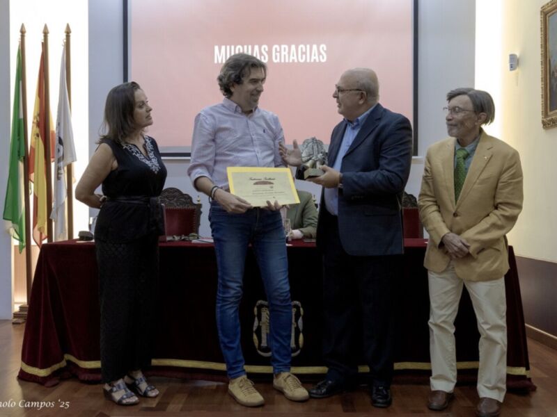 En un evento formal, una persona sostiene un diploma mientras otros observan. La escena se desarrolla en un entorno con banderas y una pantalla que dice "MUCHAS GRACIAS". La imagen transmite un ambiente de reconocimiento y agradecimiento.