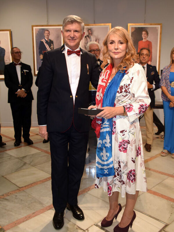 Una pareja formal posa frente a un fondo de retratos. La mujer lleva un vestido floral y el hombre una chaqueta negra con corbata roja. Ambos tienen expresiones serias y están en una sala de exposiciones o galas formales.