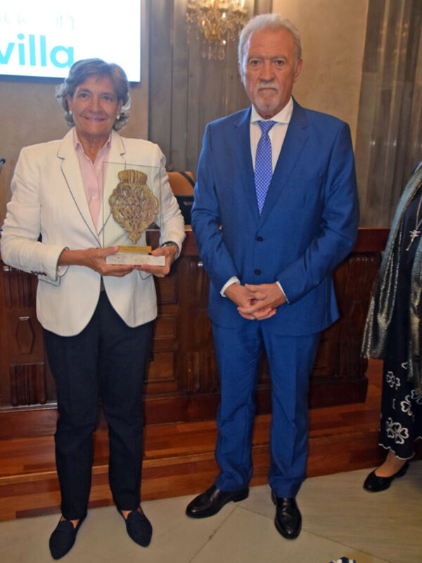 Una mujer en un traje blanco sostiene una medalla mientras está junto a un hombre en un traje azul. Ambos parecen estar en una ceremonia formal, posiblemente un evento de premiación.