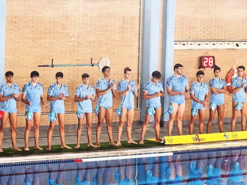 Un grupo de jóvenes nadadores en trajes de baño azul se preparan para una competencia. El fondo es un muro de ladrillos y una pared con un número 28 en la parte superior. La piscina está vacía, y las botas de natación están alineadas en el borde.