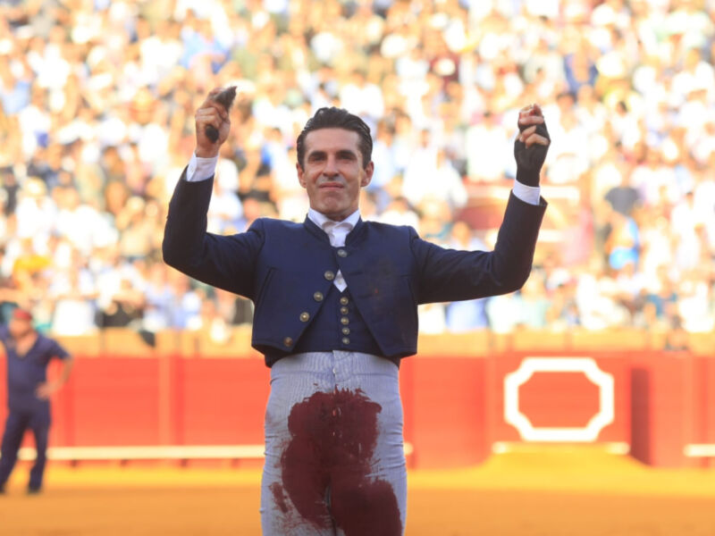 Un hombre en un traje azul y falda con una mancha roja, levanta los puños en el aire mientras un público grande lo observa.