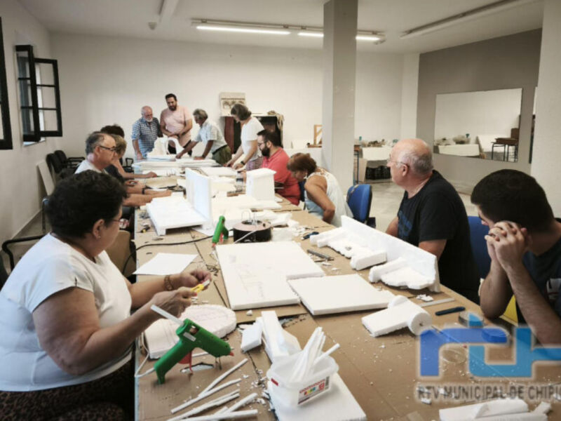 En una oficina moderna, varios empleados están trabajando en proyectos de construcción con herramientas y materiales. La imagen muestra una mesa desordenada con cajas, cartón y otros objetos de trabajo. La iluminación es buena, lo que permite ver claramente a los trabajadores y sus actividades.