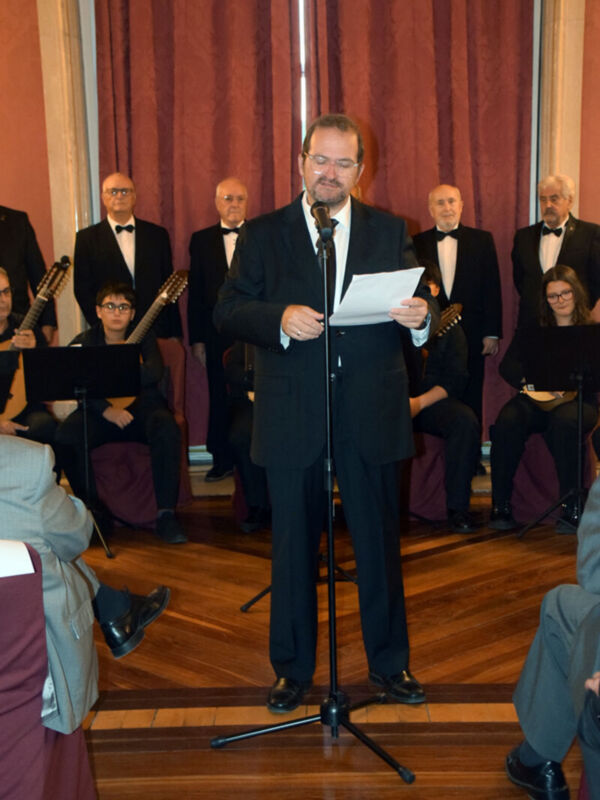 Un hombre en traje negro y corbata se encuentra en un escenario con un micrófono, sosteniendo una hoja de papel. A su alrededor, otros hombres también vestidos con trajes negros y corbatas están sentados detrás de él, algunos tocando instrumentos musicales. El fondo está decorado con cortinas rojas y un suelo de madera, creando una atmósfera formal.