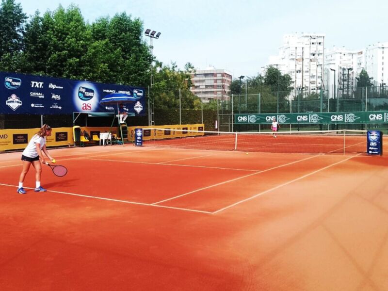 Una jugadora de tenis en un campo de tenis con una cancha roja y azul, con un cartel de publicidad en el fondo. La imagen muestra a la jugadora preparándose para un juego, con su raqueta en mano.