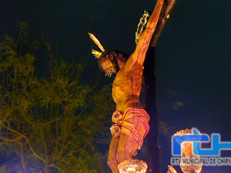 Una escultura de Jesucristo crucificado iluminada por luces doradas, con un fondo nocturno y árboles en la distancia. La escultura está en una cruz alta, con detalles como el rostro y las manos visibles. La iluminación crea un ambiente místico y dramático, destacando la figura de Jesucristo.