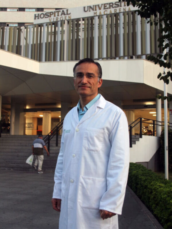 Un hombre en una camisa azul y un chaleco blanco, posando frente a la fachada de un hospital universitario. La imagen muestra una entrada principal con columnas y ventanas, junto a un par de escaleras.