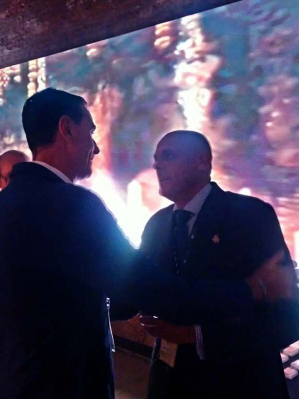 Dos hombres en trajes se saludan frente a una pantalla de fondo con luces brillantes.